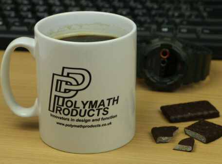 White ceramic mug with the Polymath Products logo in black print, with the tagline 'Innovators in design and function', and website address www.polymathproducts.co.uk The mug is on a desk next to some chocolate Kendal mint cake.