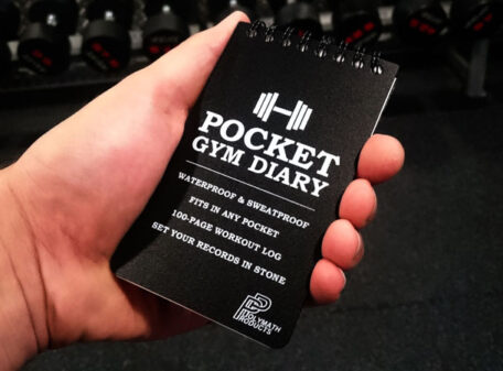 The Pocket Gym Diary held in the palm of a hand, and rack of dumbbell weights in the background.