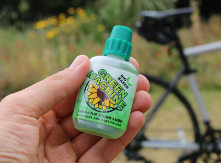 The Green Machine thin-film chain lube bottle held in the hand in a park, with a bicycle in the background.