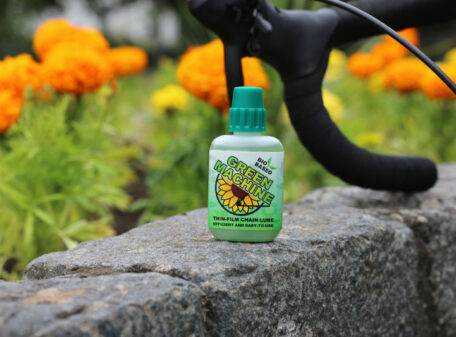 The Green Machine thin-film chain lube bottle upright on a wall in a park, with bicycle handlebars in the background.