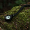 The NV-20 Compass Necklace laid on moss-covered tree branch in a woodland. Both the pendant and the neck cord are black in colour.